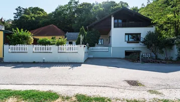 Hausfassade mit weißem Zaun und Einfahrt, umgeben von Bäumen und Grün unter blauem Himmel.