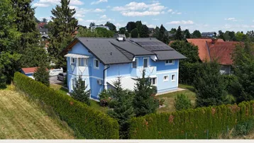 Blaues Einfamilienhaus mit Satteldach, Photovoltaikanlage und umgebender Hecke.