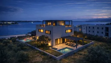 Moderne Villa am Meer bei Sonnenuntergang mit beleuchtetem Außenbereich und Pools.