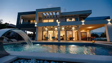 Moderne Villa bei Nacht mit beleuchtetem Pool und Wasserspiel, umgeben von einer stilvollen Terrasse.