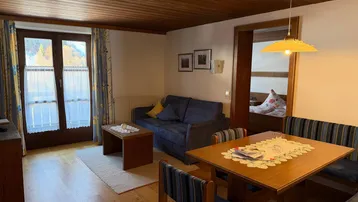 Gemütlicher Wohn-Essbereich mit Holzbalkendecke, Sofa und Esstisch, mit Zugang zum Balkon und Blick ins Grüne.