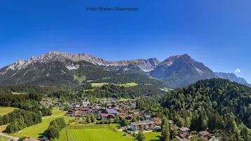 Panoramablick auf ein malerisches Dorf in den Bergen mit weiten grünen Landschaften unter blauem Himmel.