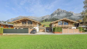 Zwei moderne Chalets mit Holzbalkonen und beeindruckendem Bergblick unter blauem Himmel.