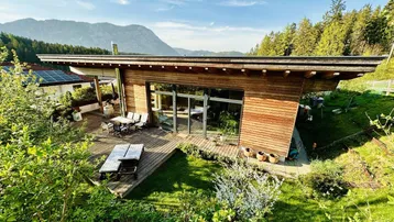 Modernes Holzhaus mit großer Terrasse, Garten und Bergblick unter blauem Himmel.