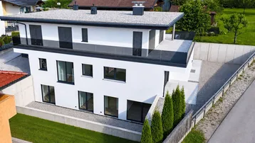 Modernes weißes Haus mit Flachdach, großen Fenstern und Balkonen in grüner Umgebung.