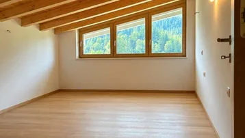 Leerer Raum mit Holzbalkendecke, Holzboden und Fenstern mit Blick ins Grüne.