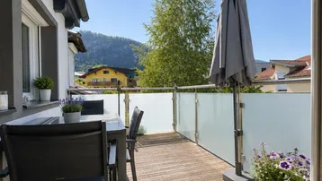 Geräumiger Balkon mit Holzboden, Glasbrüstung und Gartenmöbeln unter blauem Himmel.