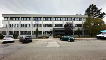 Modernes Bürogebäude mit mehreren Etagen und zahlreichen Fenstern, umgeben von Parkplätzen und Bäumen.