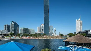 Panoramablick auf die moderne Stadtlandschaft mit einem markanten Wolkenkratzer und einem Flussufer.