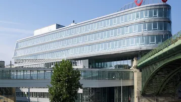 Modernes Bürogebäude mit geschwungener Glasfassade und angrenzender Brückenkonstruktion unter blauem Himmel.