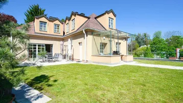 Rückansicht des Hauses mit großem Rasenbereich, Terrasse und Wintergarten unter blauem Himmel.