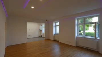 Helles Wohnzimmer mit Holzboden, Blick ins Grüne und moderner Deckenbeleuchtung mit Farbakzenten.