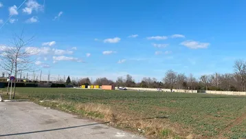 Weites, unbebautes Feld unter blauem Himmel mit einer Straße im Vordergrund und Bäumen in der Ferne.