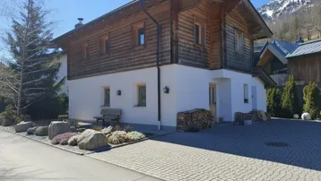 Traditionelles Holzhaus mit weißem Sockel und gepflastertem Vorplatz in alpiner Umgebung.