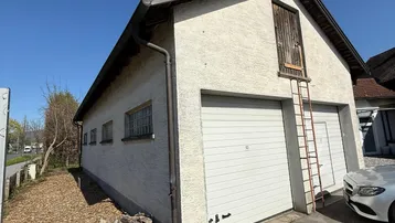 Gebäudeaußenansicht mit zwei Garagentoren, einer Leiter und einem oberen Holztor unter klarem Himmel.