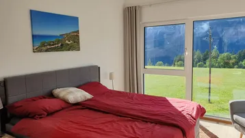 Helles Schlafzimmer mit großem Bett, roten Textilien und bodentiefen Fenstern mit Bergblick.