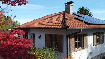 Hausfassade mit Ziegeldach, Solarpaneelen und roten Herbstbäumen im Vordergrund.