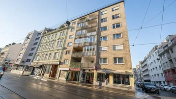 Weitwinkelaufnahme eines Mehrfamilienhauses mit Balkonen und Geschäften im Erdgeschoss an einer belebten Straße.