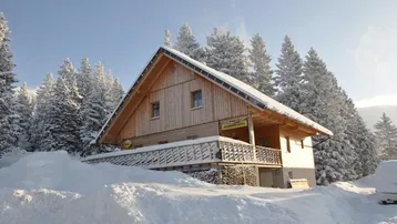 Holzhütte im Schnee mit verschneiten Bäumen und sonnigem Himmel, umgeben von einer winterlichen Berglandschaft.