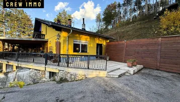 Gelbes Einfamilienhaus mit großer Terrasse und Blick auf die bewaldete Umgebung.