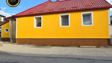 Seitenansicht eines Hauses mit leuchtend gelber Fassade und rotem Ziegeldach unter blauem Himmel.
