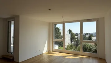 Helles, leeres Wohnzimmer mit Holzboden und großen Fenstern, die viel Tageslicht hereinlassen.