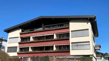 Mehrfamilienhaus mit mehreren Balkonen und Jalousien unter blauem Himmel.