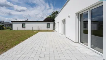 Moderne weiße Fassade mit großer gepflasterter Terrasse und angrenzendem Rasenbereich unter blauem Himmel.