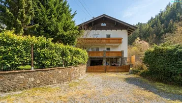 Traditionelles Haus mit Holzbalkonen, umgeben von Bäumen und einer steinernen Mauer.