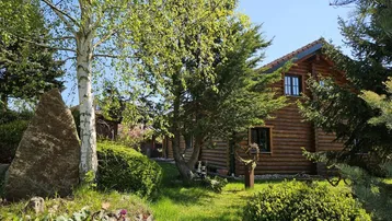 Ein charmantes Blockhaus umgeben von einem gepflegten Garten mit Bäumen und einem großen dekorativen Stein an einem sonnigen Tag.