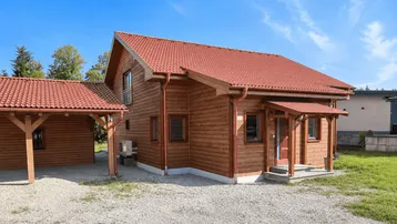 Neues Holzhaus mit rotem Ziegeldach, Kiesauffahrt und Carport unter blauem Himmel.