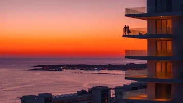 Sonnenuntergang über dem Meer mit einem modernen Gebäude und Paaren, die den Balkonblick genießen.