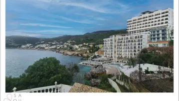 Panoramablick auf den modernen Gebäudekomplex am Meer mit felsiger Küste und bewaldeten Hügeln.