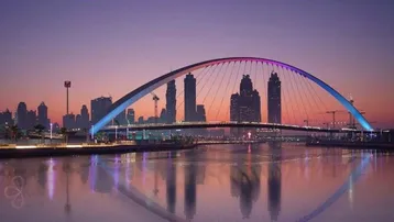 Stadtansicht bei Sonnenuntergang mit einer beleuchteten Bogenbrücke und Hochhäusern, die sich im Wasser spiegeln.