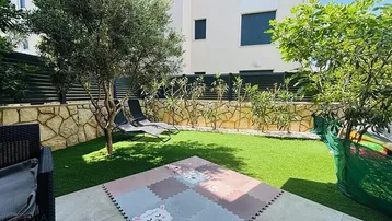 Gepflegter Gartenbereich mit Kunstrasen, Olivenbaum und Liegestühlen, ideal zum Entspannen und Spielen.