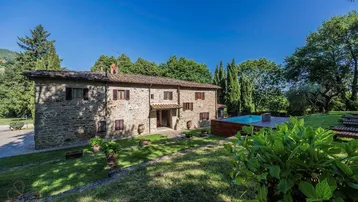 Traditionelles Steinhaus mit weitläufigem Garten, Pool und üppiger Vegetation unter blauem Himmel.