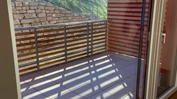 Heller Blick vom Innenraum auf einen sonnigen Balkon mit Holzelementen und grüner Hügellandschaft.