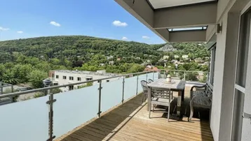 Langer Holzbalkon mit Sitzgelegenheiten und weitem Blick auf die grüne Landschaft und umliegende Häuser.
