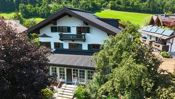 Traditionelles Haus mit Satteldach und grüner Umgebung, umgeben von Bäumen und Wiesen.
