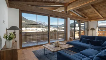 Gemütliches Wohnzimmer mit Holzbalkendecke, großen Fenstern und herrlichem Bergblick auf die verschneite Landschaft.