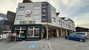 Modernes Geschäftsgebäude mit mehreren Etagen, Geschäften im Erdgeschoss und Parkplätzen davor.
