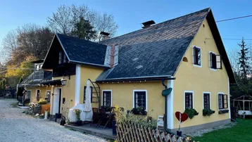 Gelbes Landhaus mit dunklem Dach, kleinen Fenstern und einer kleinen Terrasse im Freien.