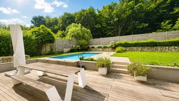 Große Holzterrasse mit Esstisch, Pool und weitläufigem Gartenbereich unter blauem Himmel.