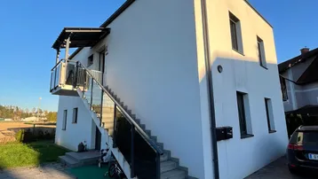 Modernes weißes Gebäude mit externer Treppe und gepflastertem Vorplatz unter blauem Himmel.