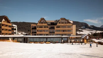 Modernes Gebäude in den Bergen mit Holzfassade und großen Fenstern, umgeben von Schnee und Skipisten.