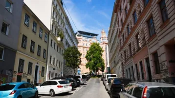 Blick auf eine belebte Stadtstraße mit geparkten Autos und hohen Wohngebäuden unter blauem Himmel.