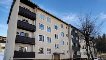 Mehrfamilienhaus mit Balkonen und dunklen Holzverkleidungen, umgeben von Schnee und Bäumen unter blauem Himmel.