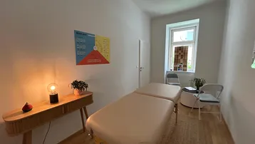 Heller Therapieraum mit Massageliege, Holztisch und Fenster, ideal für Behandlungen und Entspannung.