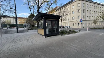 Kleiner, freistehender Kiosk auf einem gepflasterten Platz mit umliegenden Bäumen und Gebäuden.