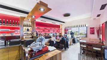 Restaurantbereich mit Gästen, Holztischen und einer Theke mit Aufschnittmaschine, die auf frische Speisen hinweist.
